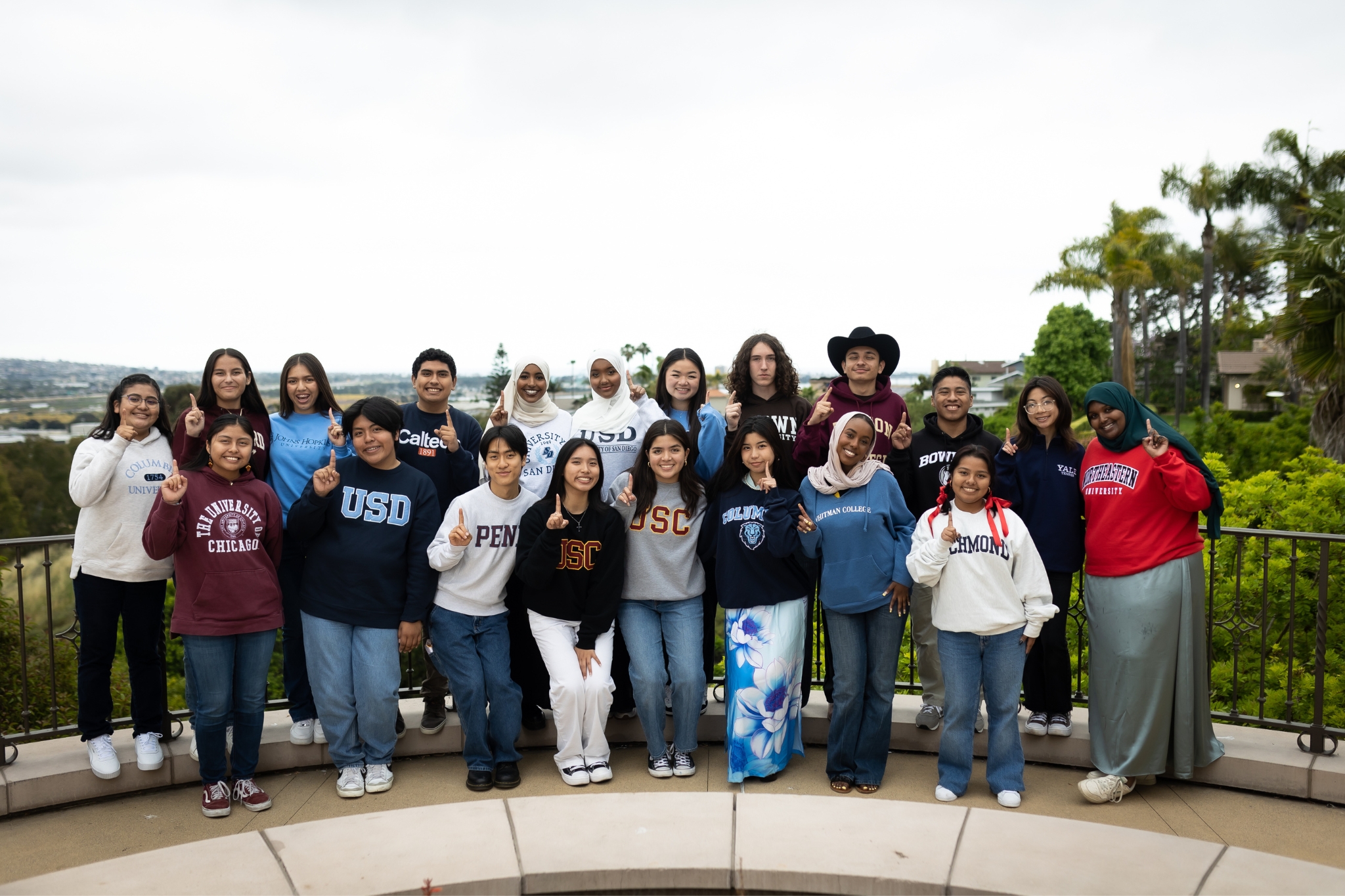 First Gen Scholars: A group of young people in college sweatshirts holding up their index fingers smile into the camera